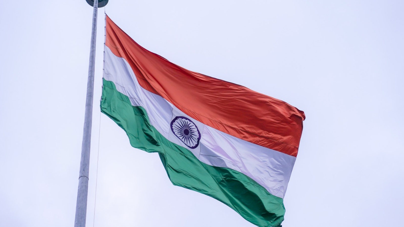 The flag of India flying from a tall flagpole against an overcast sky, the 24-spoke Ashoka Chakra at its centre