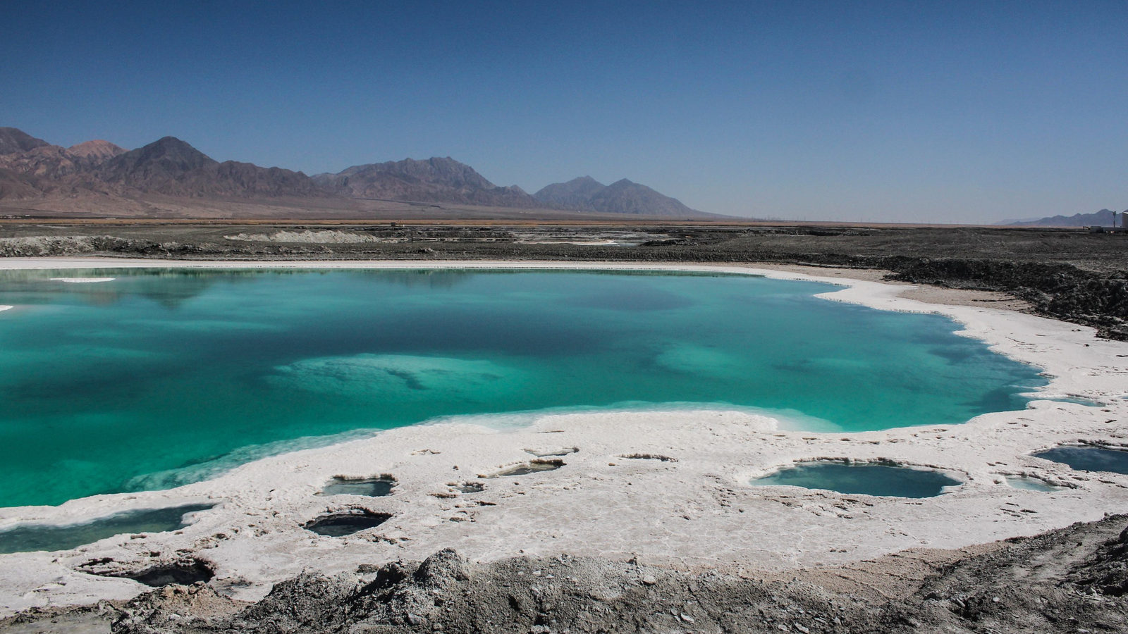 A turquoise salt lake nestled among barren, rocky volcanic mountains under a clear sky