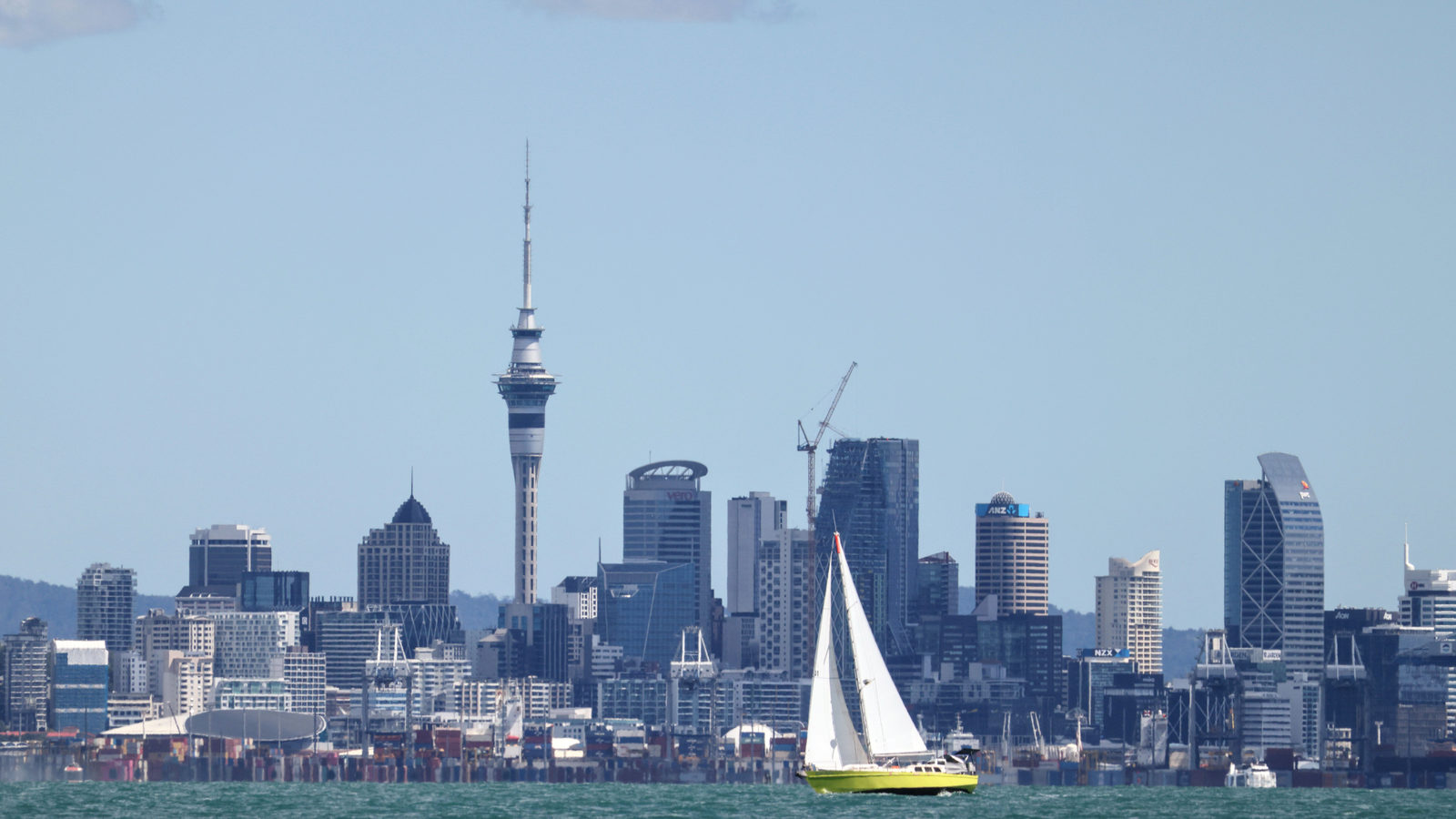 A sailboat crosses Auckland's harbour with the city skyline and Sky Tower rising behind it under a clear blue sky