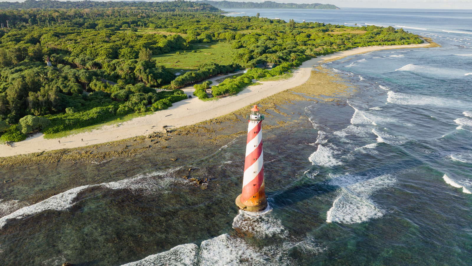 Aerial view of the red-and-white striped Indira Point Lighthouse on a forested spit of Great Nicobar Island, with waves breaking on both shores