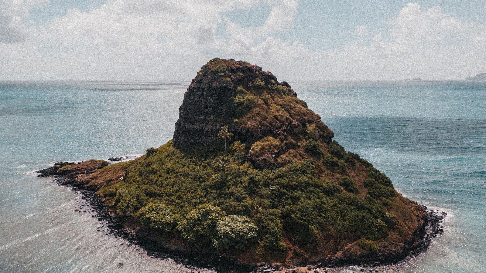 Aerial view of a small, isolated rocky green island surrounded by open turquoise ocean
