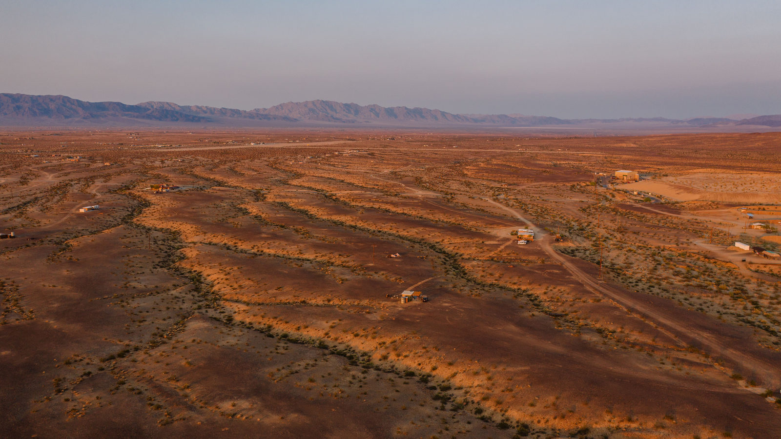 Aerial view of a vast, sunlit orange desert landscape with faint mountains on the horizon