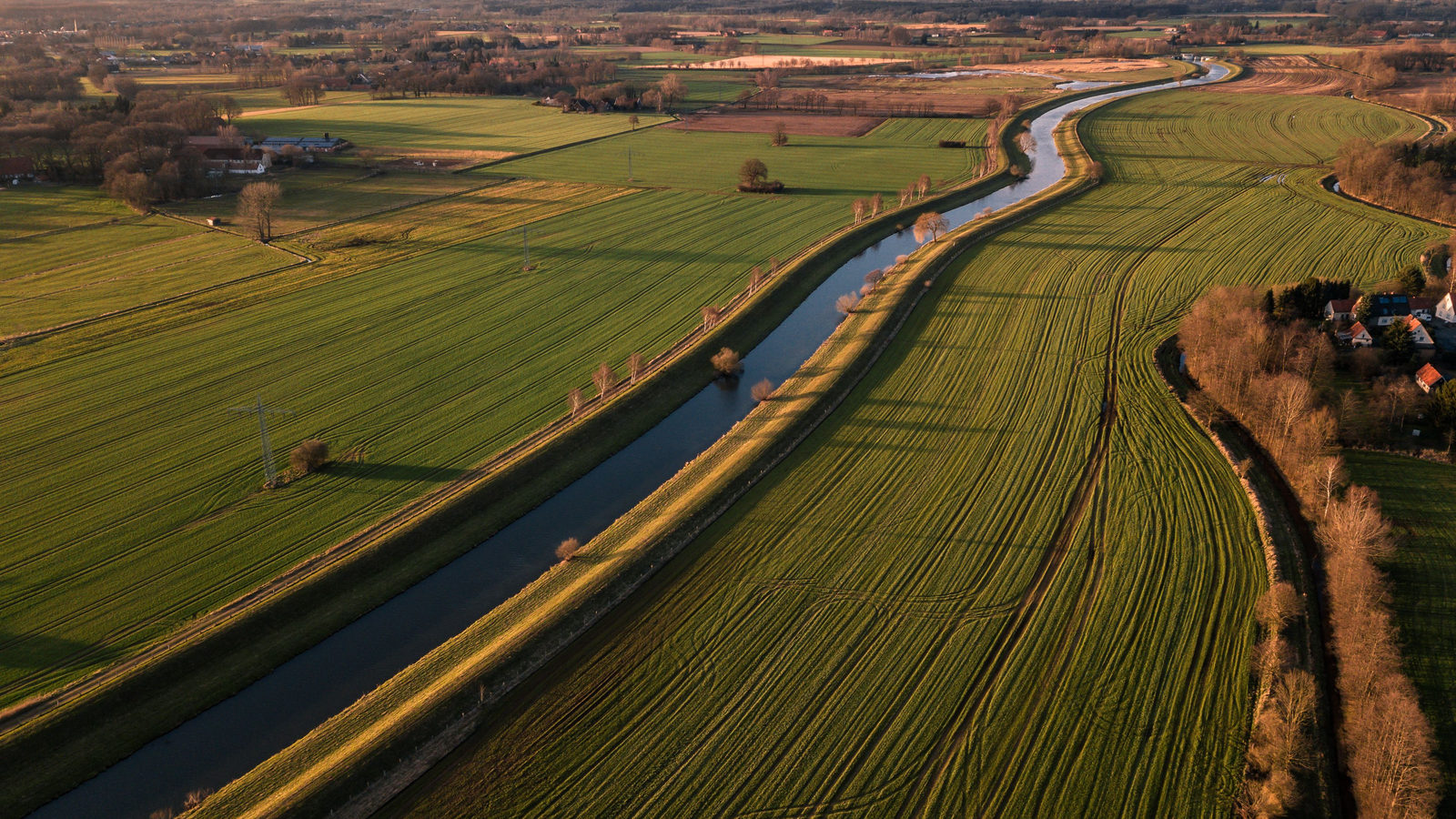 An aerial view of a straight drainage canal running through flat, geometric green farmland in the Dutch countryside