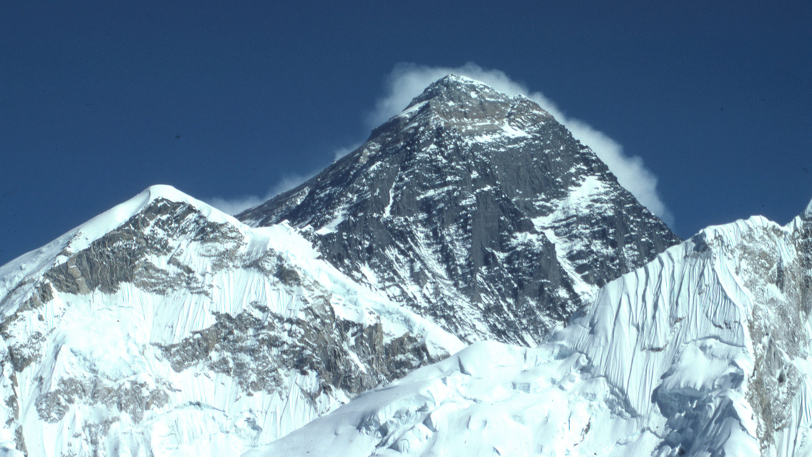 The snow-covered summit pyramid of Mount Everest rising above surrounding peaks under a dark blue sky, with wind-blown snow trailing from the top