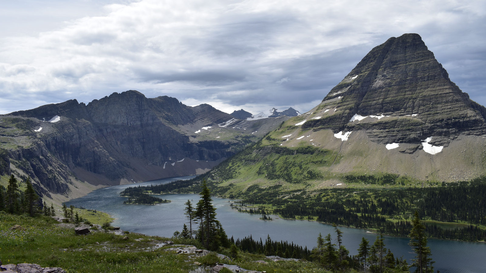 A turquoise alpine lake surrounded by forest beneath a jagged, pyramid-shaped mountain peak in Glacier National Park, Montana