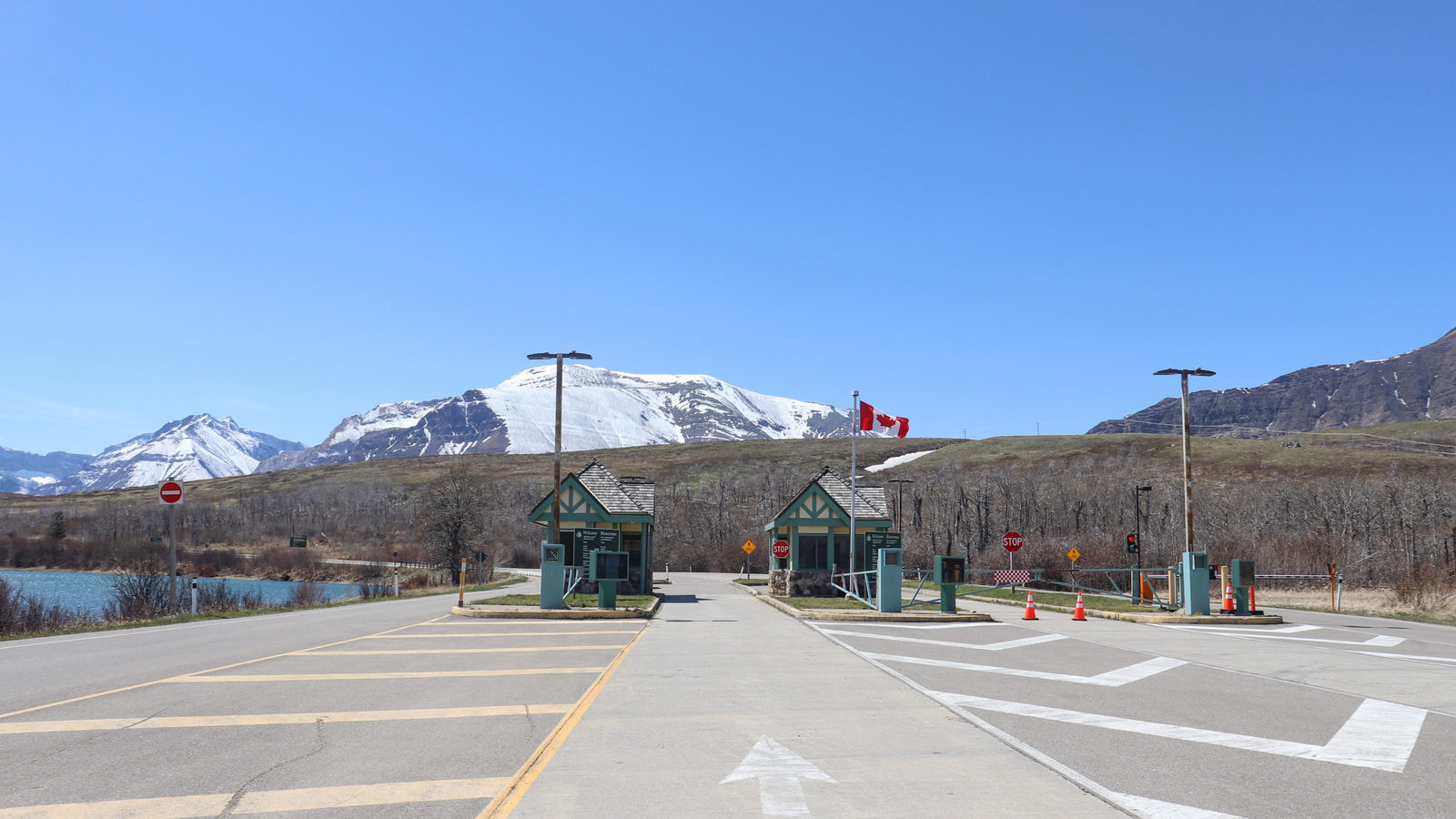 A car travels down a highway lane toward a US-Canada border checkpoint with inspection booths, flags, and snow-capped mountains in the distance