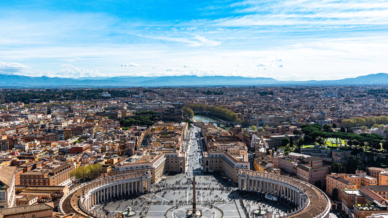 An aerial view of St. Peter's Square and Basilica in Vatican City, with the curved colonnade and obelisk visible against the Rome skyline under a blue sky